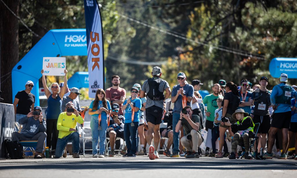 A runner crosses the finish line of the Kodiak Ultra Marathons in Big Bear, surrounded by cheering crowds and race banners.