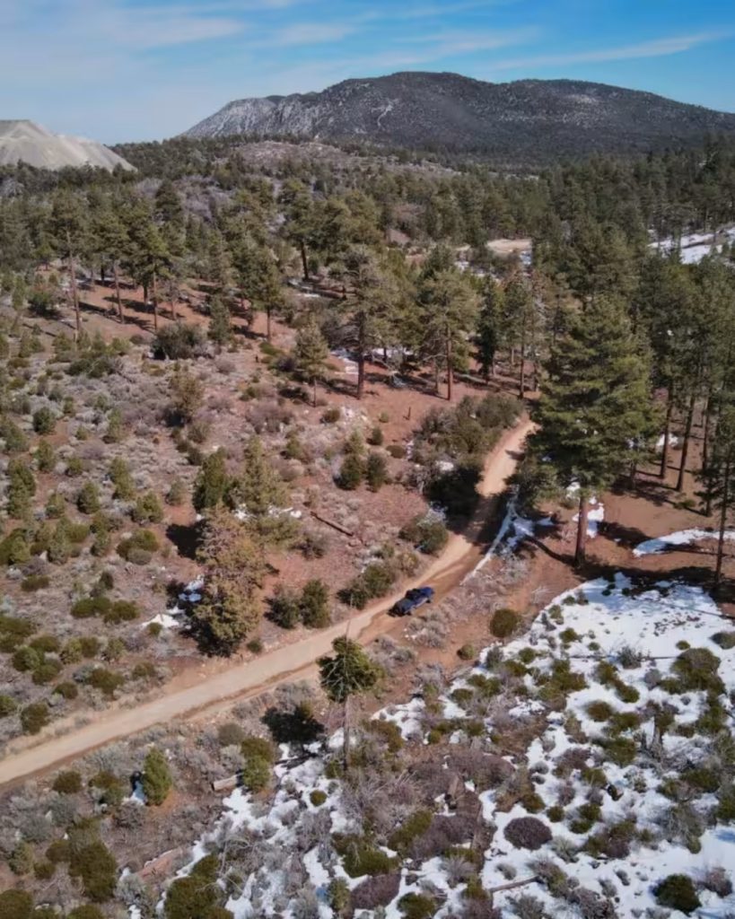 Aerial view of Holcomb Valley Road (3N16) winding through pine forests with patches of snow and scenic mountain landscapes in Big Bear.