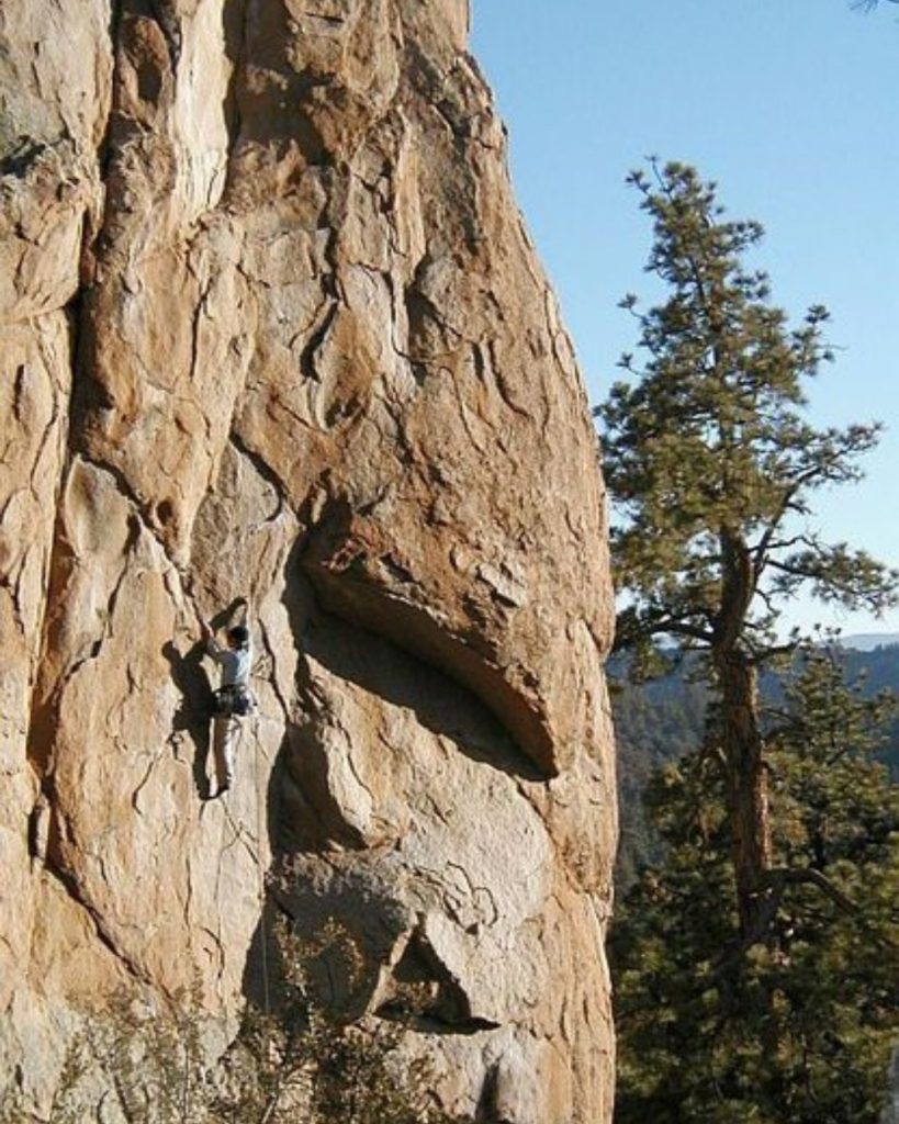 A rock climber ascends a towering granite wall at Holcomb Valley surrounded by scenic mountain forest in Big Bear.