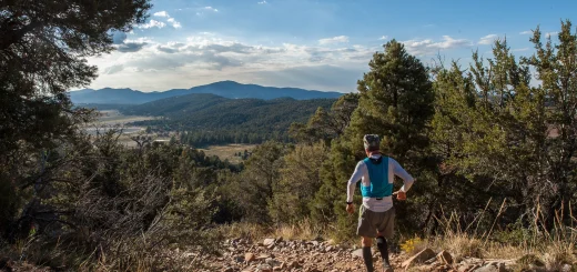 A trail runner runs down a rocky path, looking out at the Big Bear landscape with distant mountains under a cloudy blue sky.