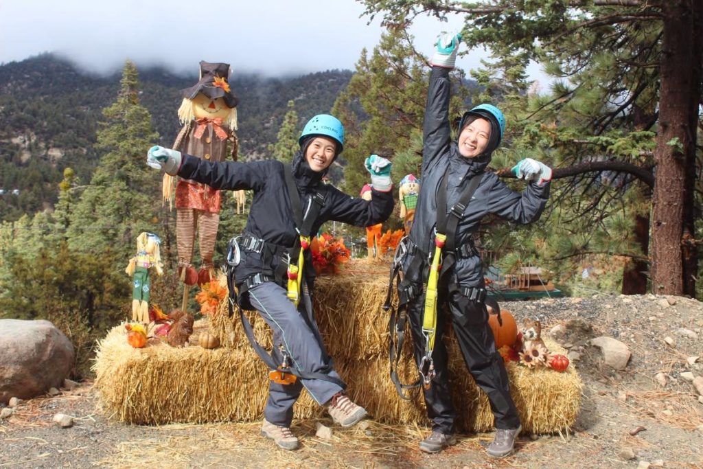 Two smiling participants wearing zipline helmets and harnesses pose beside pumpkins, hay bales, and scarecrows during a fall adventure in Big Bear Lake.
