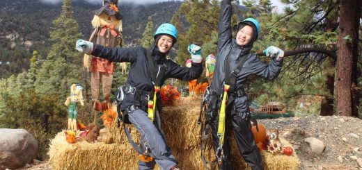 Two smiling participants wearing zipline helmets and harnesses pose beside pumpkins, hay bales, and scarecrows during a fall adventure in Big Bear Lake.