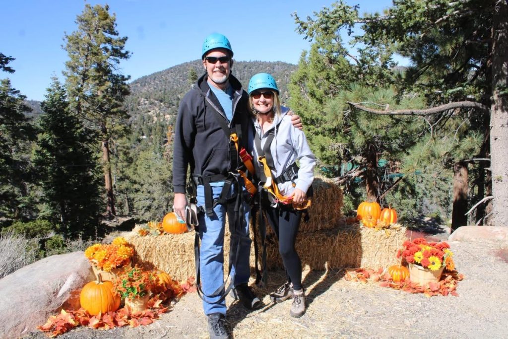 Two participants wearing blue helmets and safety harnesses pose with pumpkins and fall decorations during a zipline adventure in Big Bear Lake.