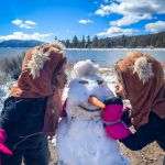 Two children in winter clothing building a snowman at Big Bear Lake, surrounded by snow-covered trees and a clear sky.