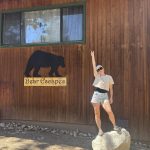 Woman posing outdoors beside a wooden cabin wall with a “Bear Escapes” sign featuring a bear silhouette, standing on a rock while pointing upward under bright sunlight.