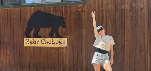Woman posing outdoors beside a wooden cabin wall with a “Bear Escapes” sign featuring a bear silhouette, standing on a rock while pointing upward under bright sunlight.