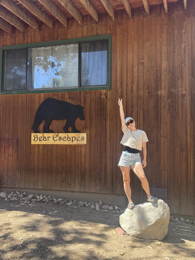 Woman posing outdoors beside a wooden cabin wall with a “Bear Escapes” sign featuring a bear silhouette, standing on a rock while pointing upward under bright sunlight.