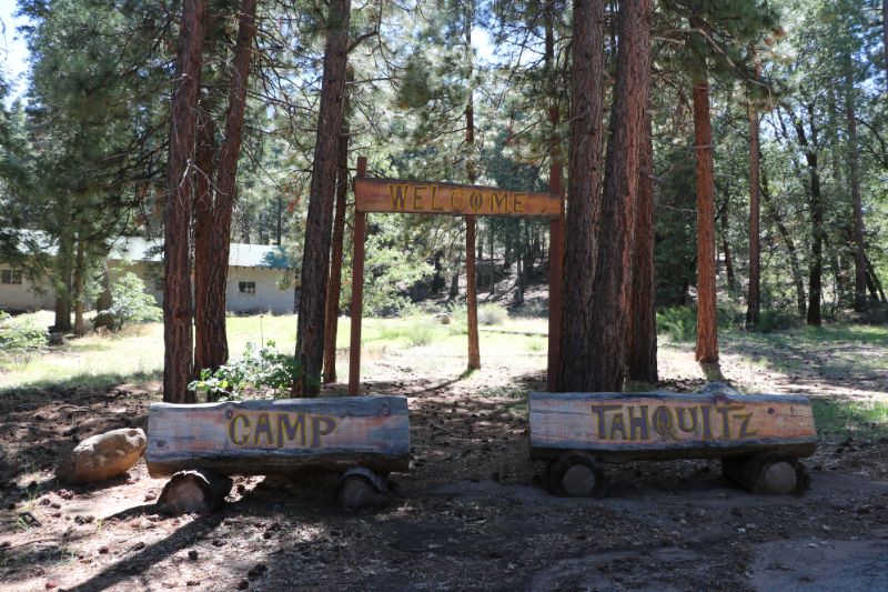 Wooden welcome sign at Camp Tahquitz surrounded by tall pine trees in a forested setting near Big Bear.