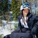 A smiling woman in a helmet and goggles sits on a ski lift, holding her snowboard as she looks down the snowy mountain at Big Bear.