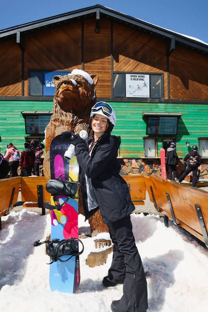 A woman wearing a helmet and goggles smiles while posing with her snowboard next to a large, carved wooden bear statue at Big Bear.