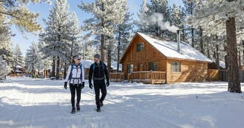 Man and woman in ski coats walking down a snowy road with a log cabin and snow covered trees in the background