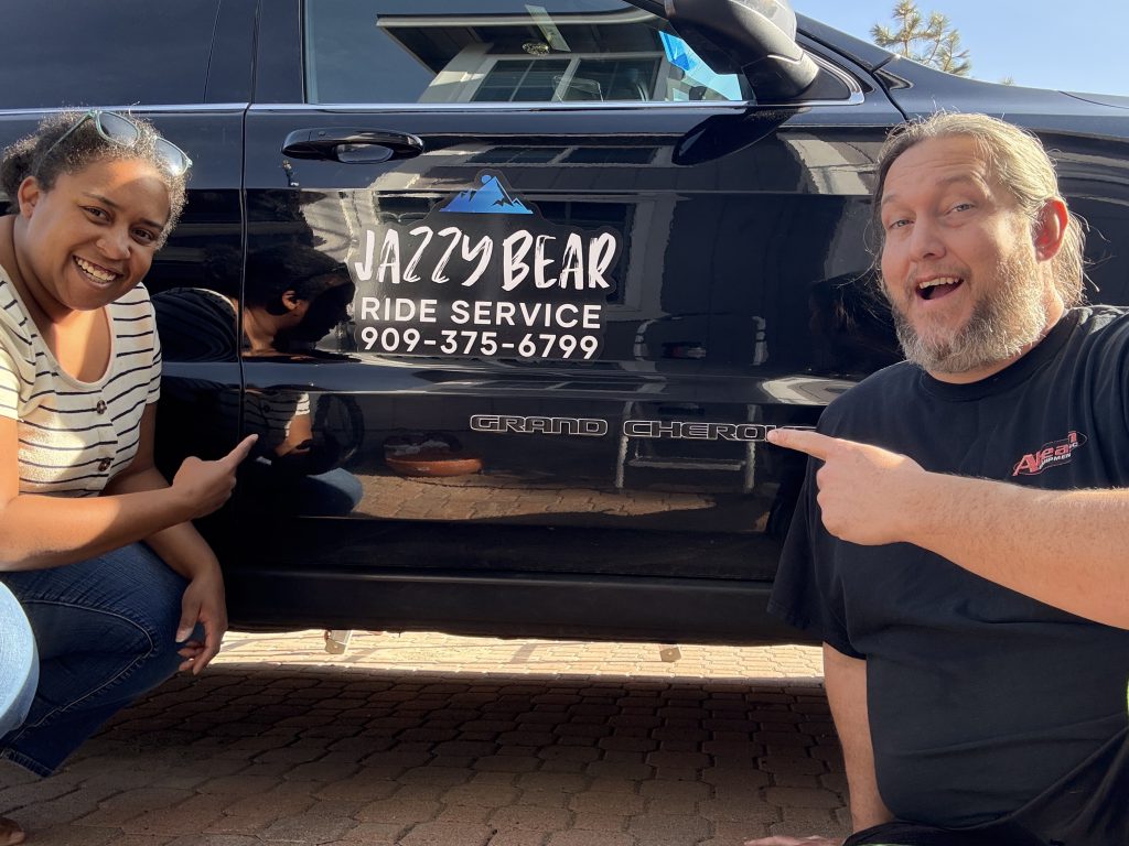 A man and woman kneel beside a black vehicle with "Jazzy Bear Ride Service" and a phone number on its side, pointing to the text, in Big Bear.
