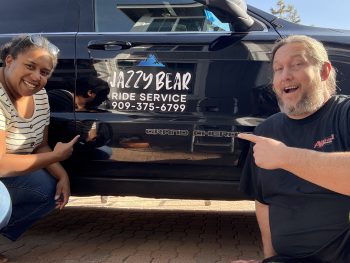 A man and woman kneel beside a black vehicle with "Jazzy Bear Ride Service" and a phone number on its side, pointing to the text, in Big Bear.
