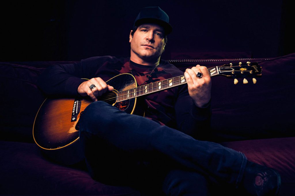 A man wearing a baseball cap and dark clothing sits on a couch, holding an acoustic guitar and looking at the camera at Big Bear