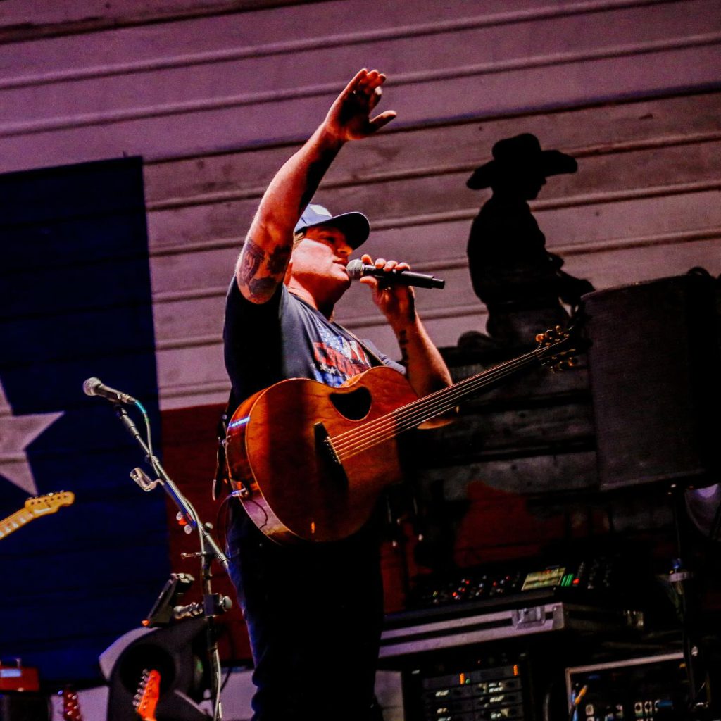 A male musician with tattoos on his arm holds a microphone and an acoustic guitar while raising his other hand, performing in front of a Texas flag at Big Bear