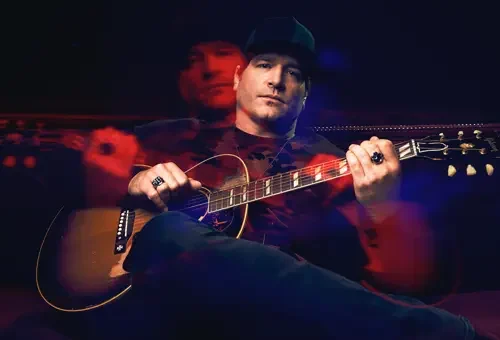 A man wearing a baseball cap and holding an acoustic guitar sits on a couch, with a red and blue light creating a blurred, double-exposure effect around him at Big Bear