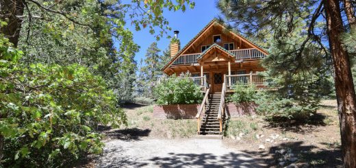 A log cabin in the mountains surrounded by pine trees and blue skies. Stairs leading up to the log cabin are on center.