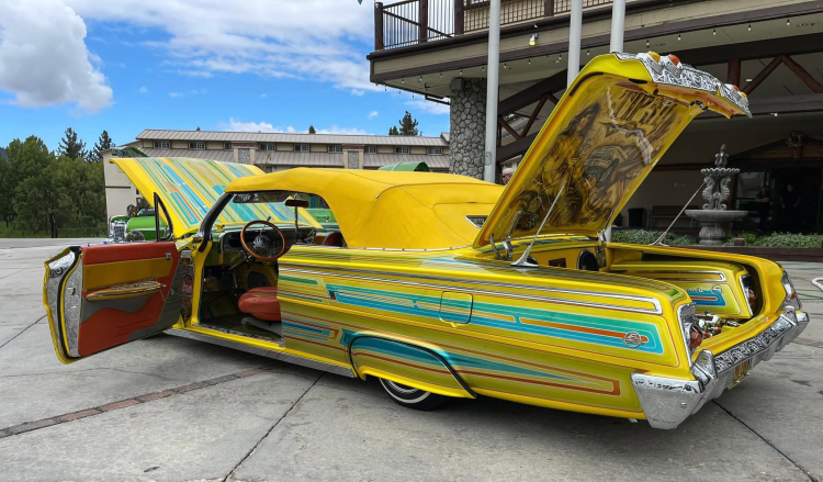 Bright yellow custom lowrider car with colorful pinstriping, open hood, trunk, and doors displayed at a car show in Big Bear Lake.