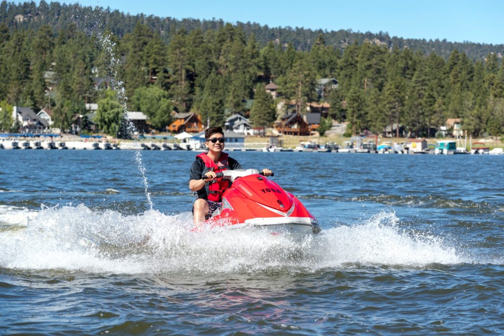 Person riding a jet ski across Big Bear Lake with mountain scenery and lakeside cabins in the background