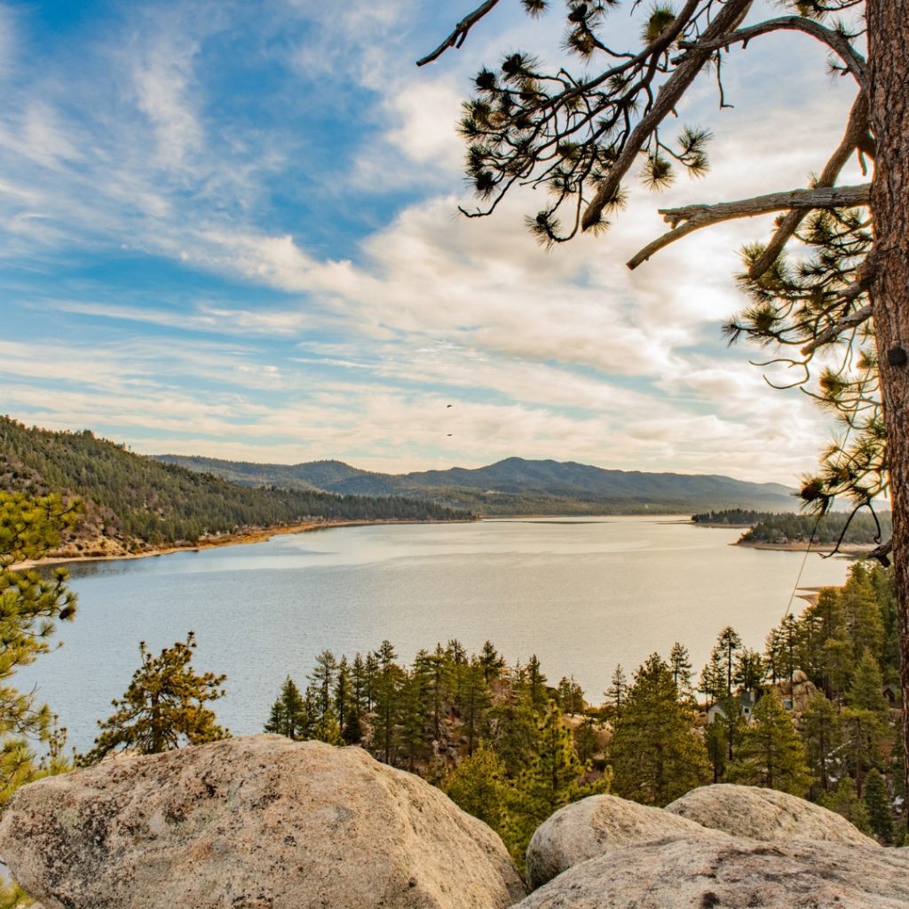 Scenic overlook of a large mountain lake surrounded by pine forests and rolling hills, viewed from rocky foreground under a partly cloudy sky with tree branches framing the top.