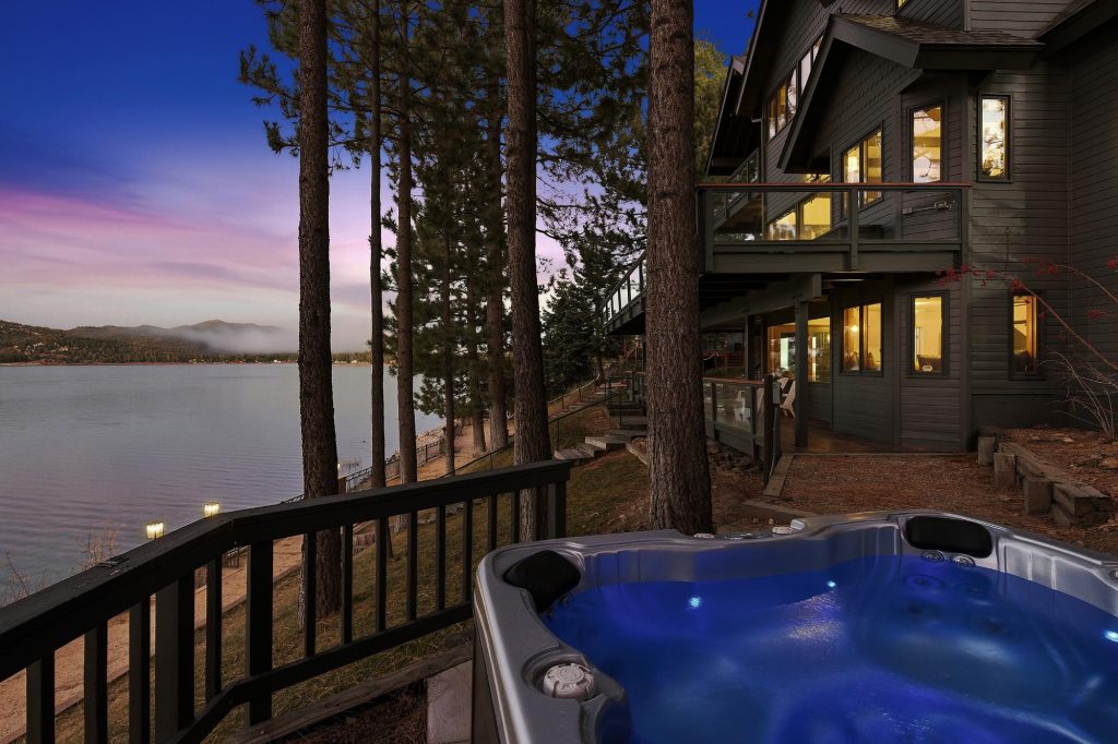 Lakefront backyard at dusk with hot tub in the foreground, pine trees, and scenic lake view