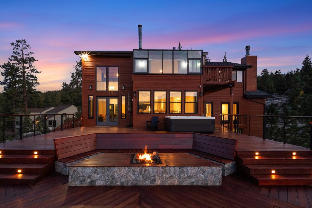 Luxury lakefront cabin exterior featuring a spacious patio, outdoor fire pit, hot tub, and mountain views at sunset