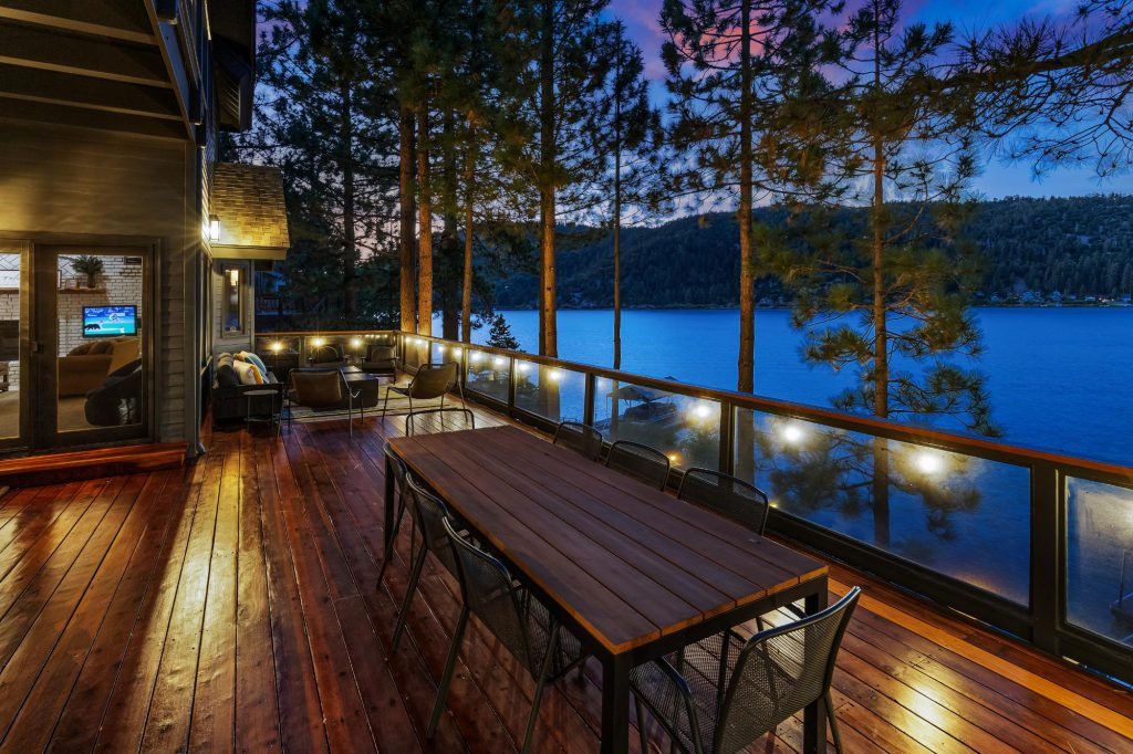 Luxury lakefront deck overlooking Big Bear Lake at dusk with outdoor dining table, seating area, pine trees, and illuminated waterfront views.