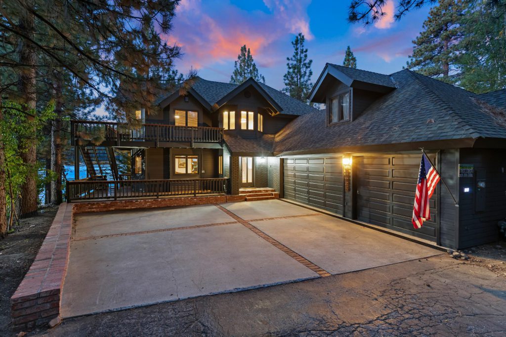A lakefront cabin in Big Bear at dusk, featuring a driveway and garage, surrounded by trees and a serene atmosphere.