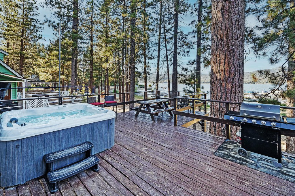 A wooden deck with a hot tub, picnic table, and barbecue grill is surrounded by tall pine trees, overlooking a lake in Big Bear.
