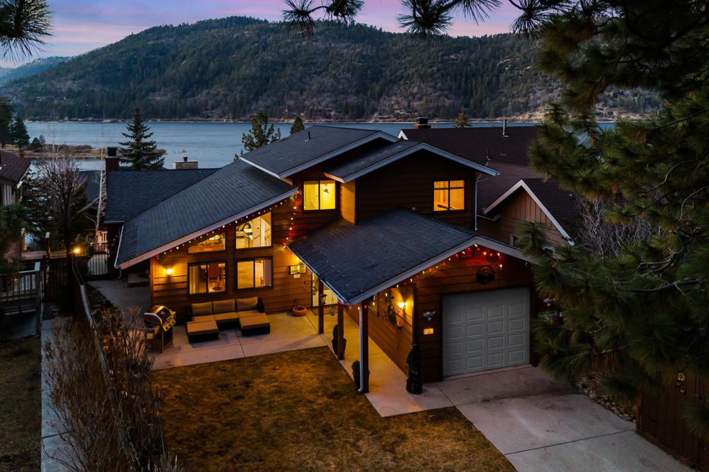 An inviting, multi-story home with lighted windows and a two-car garage is pictured at dusk, with the serene waters of Big Bear Lake and a mountain visible in the background.