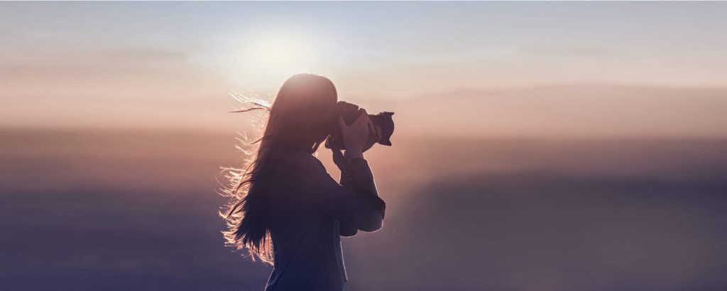 A silhouette of a woman with long hair taking a photo with a camera against a hazy, sunlit sky at Big Bear