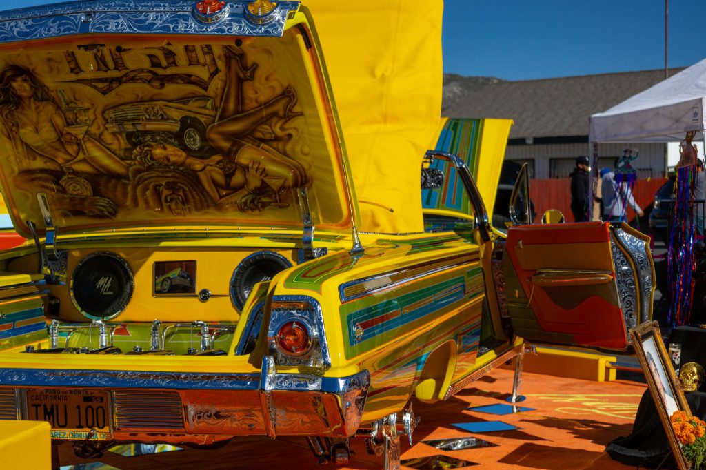 Yellow custom lowrider with airbrushed artwork and open trunk displaying speakers at a car show.
