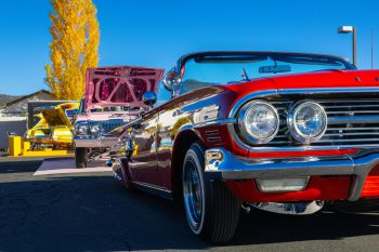 Red classic lowrider car displayed at an outdoor car show with other vintage cars lined up behind it.