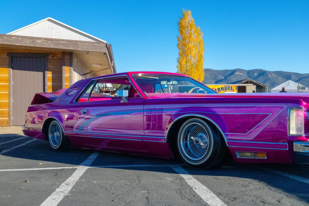 Bright pink custom lowrider parked outdoors with mountains and autumn trees in the background.