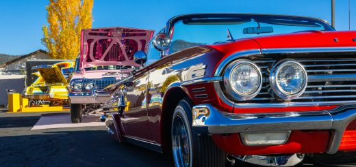 Red classic lowrider car displayed at an outdoor car show with other vintage cars lined up behind it.