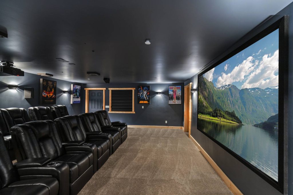 A modern home theater in Big Bear with multiple rows of black leather recliners facing a large screen displaying a scenic mountain and lake view.