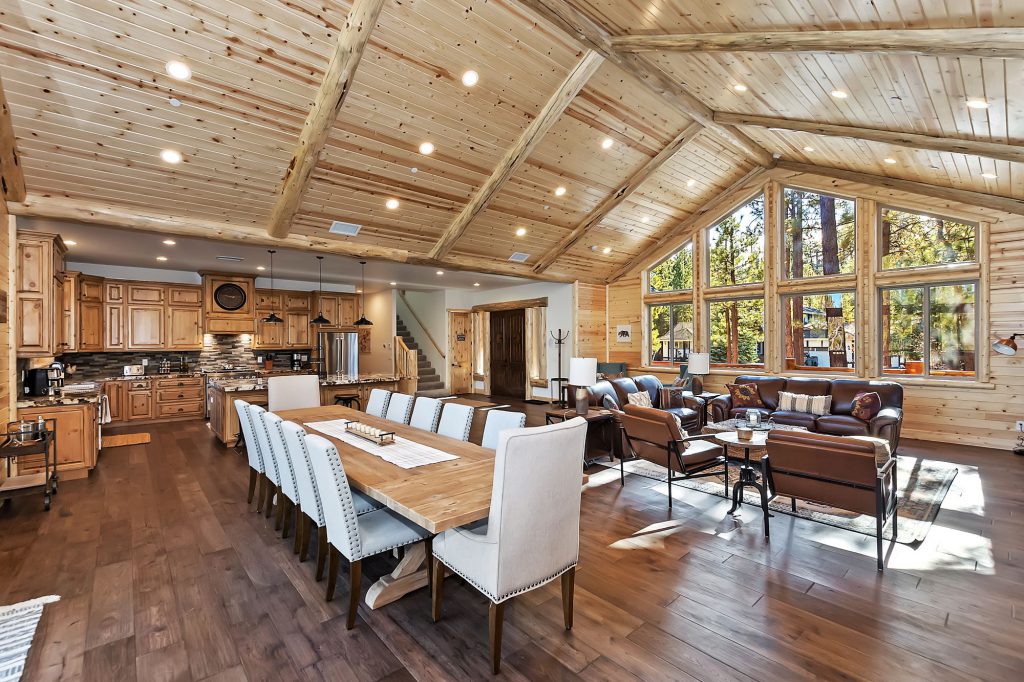 A large, open-concept living area with a long dining table, a living room, and a kitchen is visible, with high vaulted wood ceilings and large windows in Big Bear.