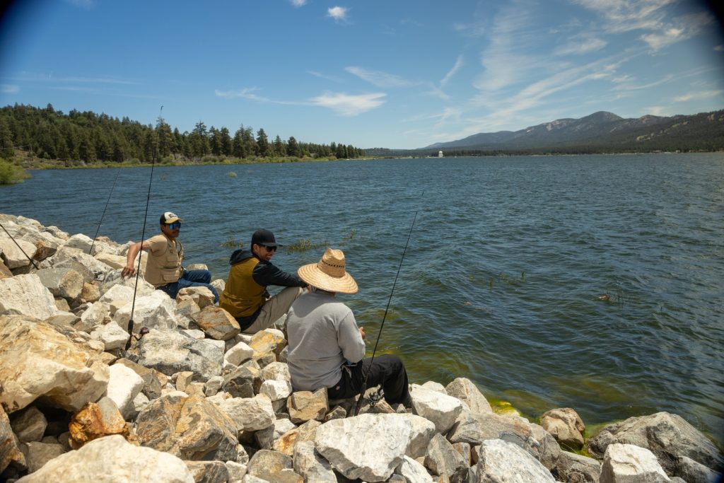 Three people fishing along the rocky shoreline of Big Bear Lake with mountains and pine trees in the background under a clear blue sky.