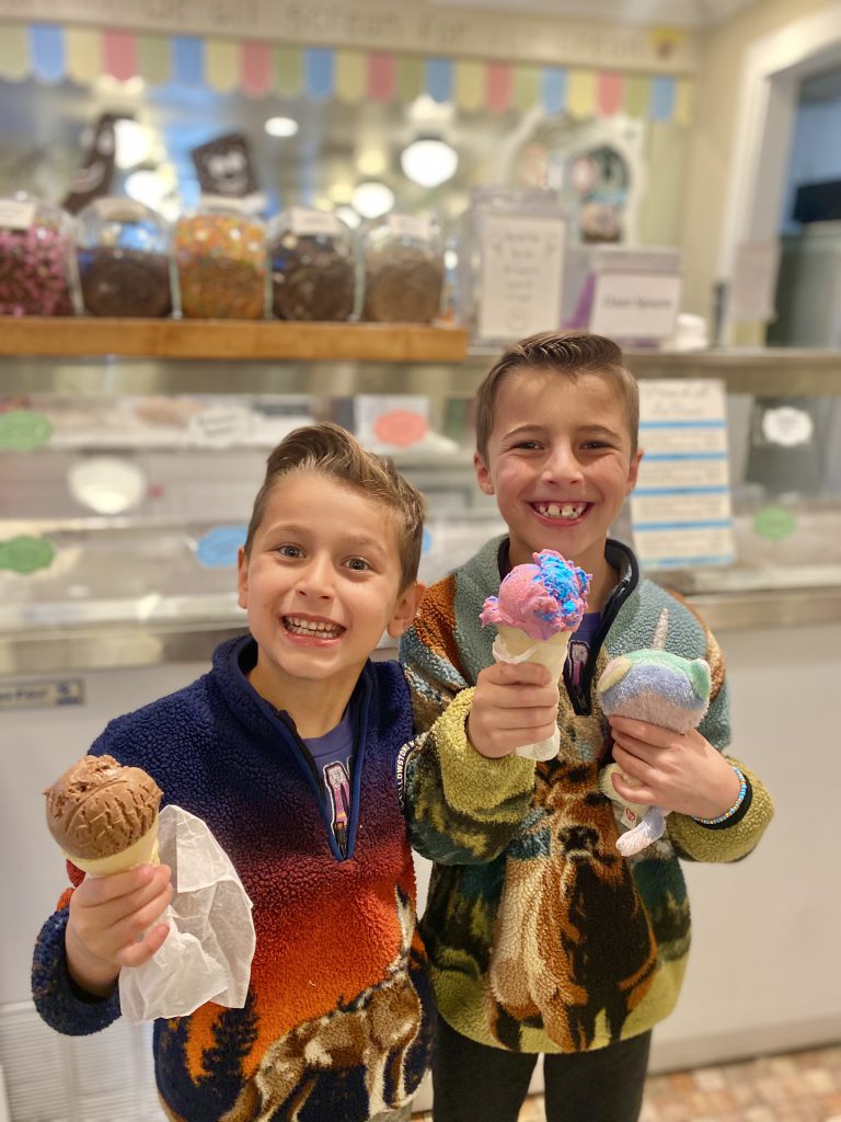 Two boys are smiling and holding ice cream cones in an ice cream shop in Big Bear, with one boy holding a chocolate cone and the other holding a multi-colored one.