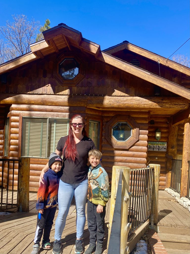 A mother smiles while standing on the porch of a log cabin at Big Bear with her two young sons, who are also smiling and looking at the camera.
