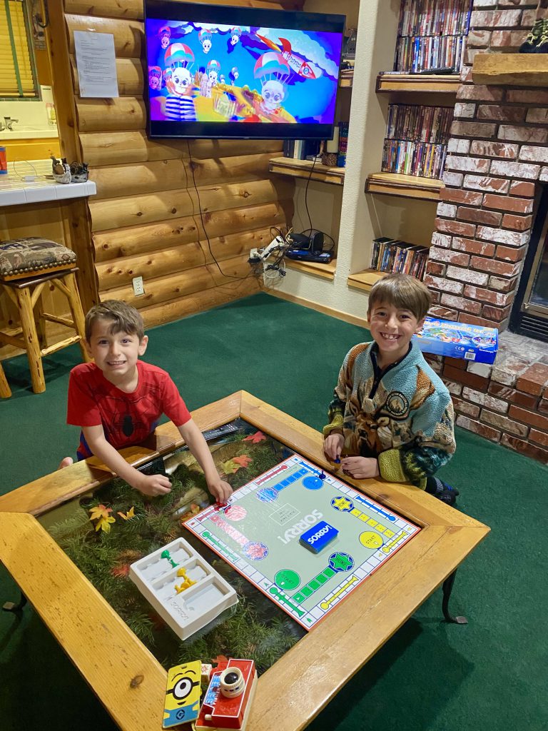 Two boys are smiling while sitting on the floor and playing a board game on a unique wooden coffee table at Big Bear, with a TV visible in the background.