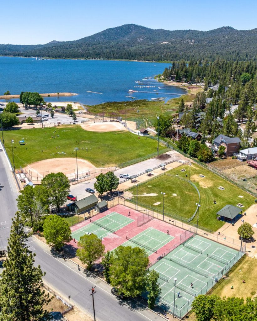 Aerial view of Meadows Park in Big Bear featuring tennis courts, baseball fields, open green spaces, and scenic Big Bear Lake in the background.