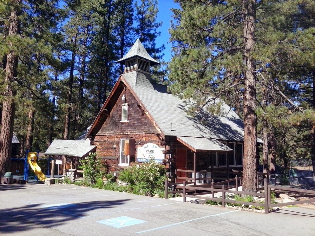Historic Miller Park building in Big Bear surrounded by tall pine trees, with nearby playground and community recreation facilities.