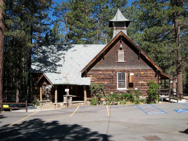 Front exterior view of the historic Miller Park building in Fawnskin featuring rustic log construction, a bell tower, and surrounding pine trees near Big Bear Lake.