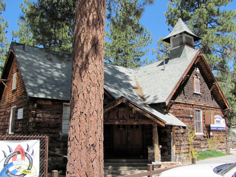 Historic Miller Park Schoolhouse in Fawnskin featuring rustic log architecture, a bell tower, and towering pine trees in the Big Bear Lake area.