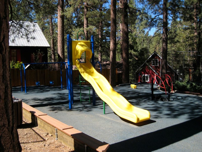 Children's playground at Miller Park in Fawnskin featuring a bright yellow slide, swings, and play structures surrounded by towering pine trees near Big Bear Lake.
