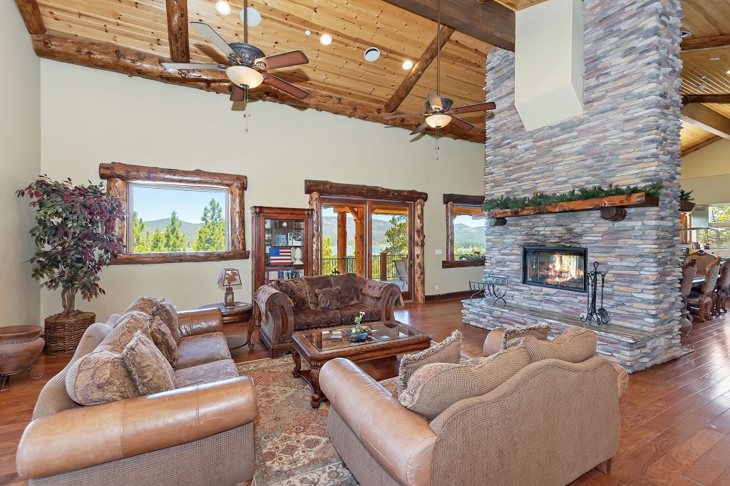 A large, rustic living room with brown couches and a tall, multi-sided stone fireplace in the center of the room in Big Bear.