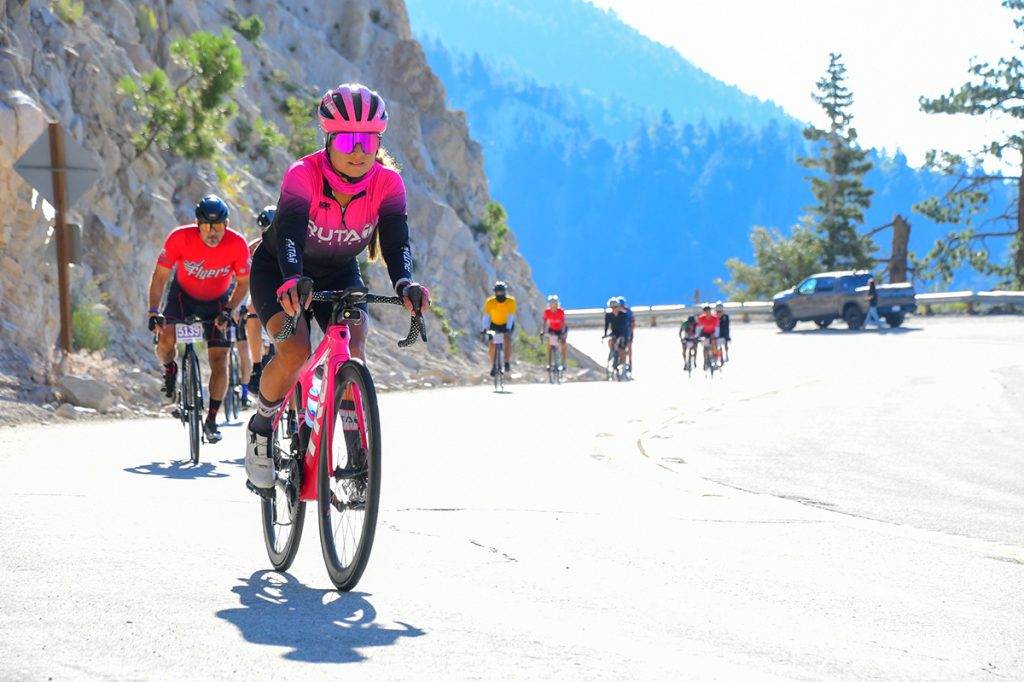 Group of cyclists competing in a road cycling race on a mountain highway with rocky cliffs, clear blue sky, and a lead rider in a pink jersey.