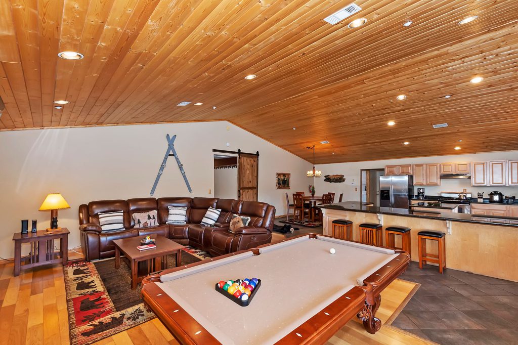 A large open-concept room with a pool table in the foreground, a brown leather sectional couch with a rug, and a kitchen with bar seating in the background, all under a wood-paneled ceiling in a Big Bear cabin.