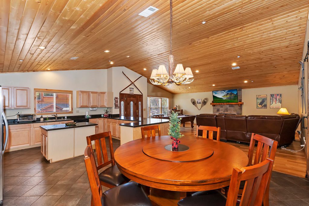 A large open-concept room with a round dining table in the foreground, leading to a kitchen with wooden cabinets and a living room with a stone fireplace and leather couches, all under a vaulted wood ceiling in a cabin in Big Bear.
